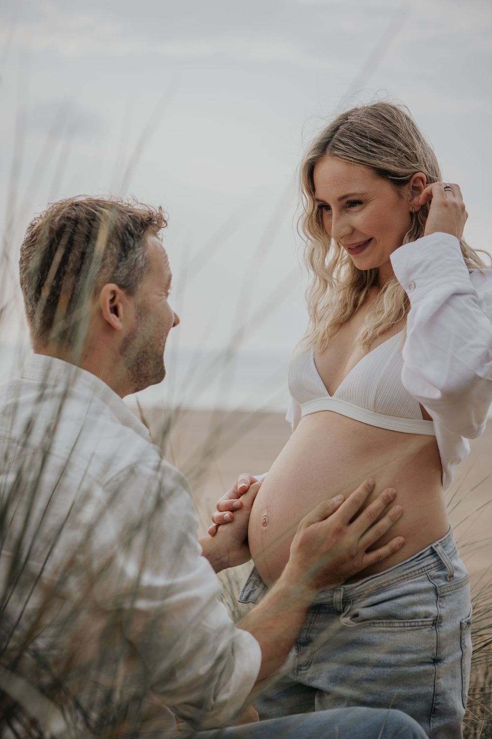 Man kneeling beside pregnant woman in white bralette and jeans, gently touching her baby bump outdoors. Cornwall Maternity Photographer