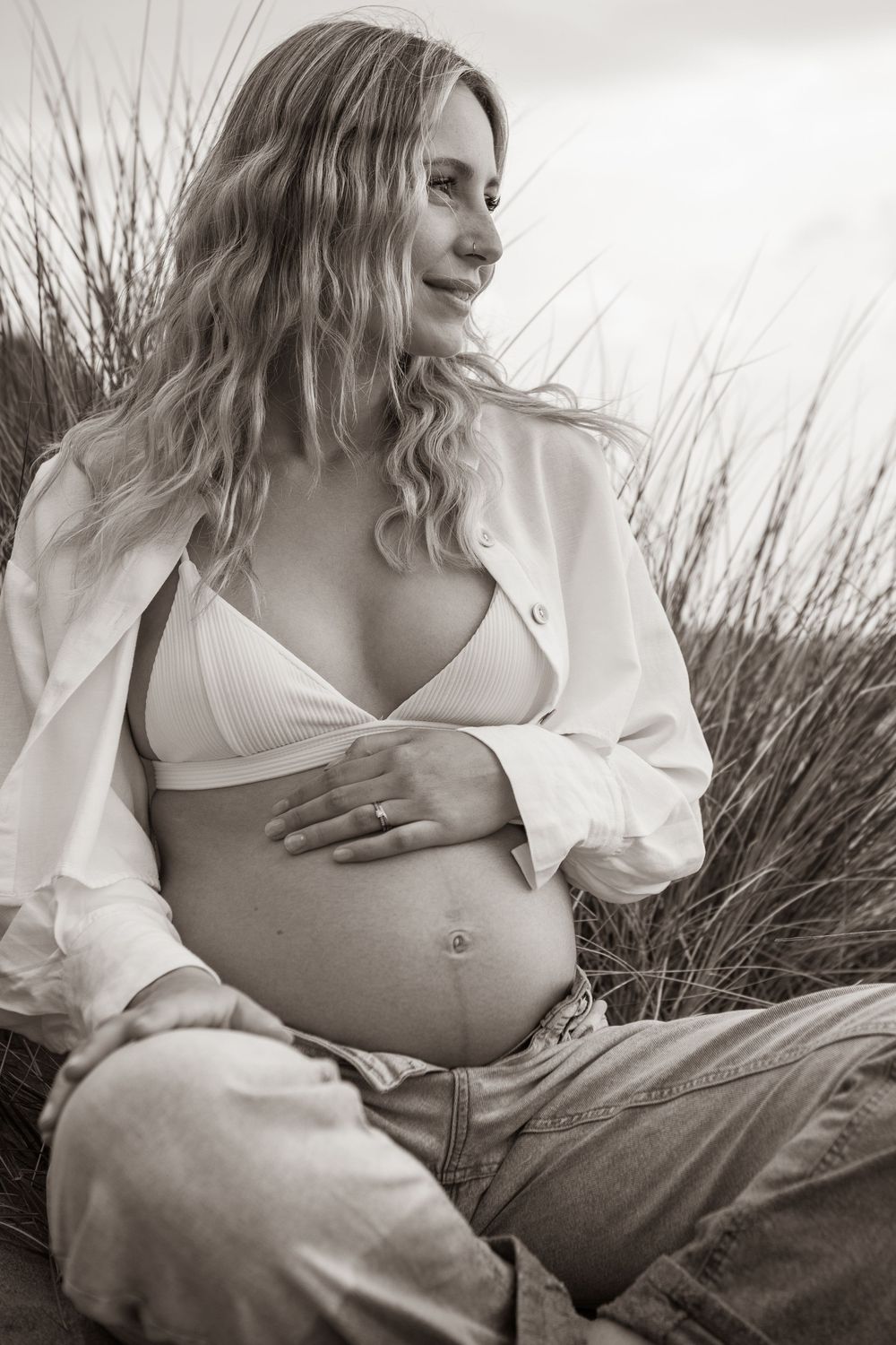 Black and white portrait of pregnant woman in white bralette and open shirt sitting in tall grass. Cornwall Maternity Photographer