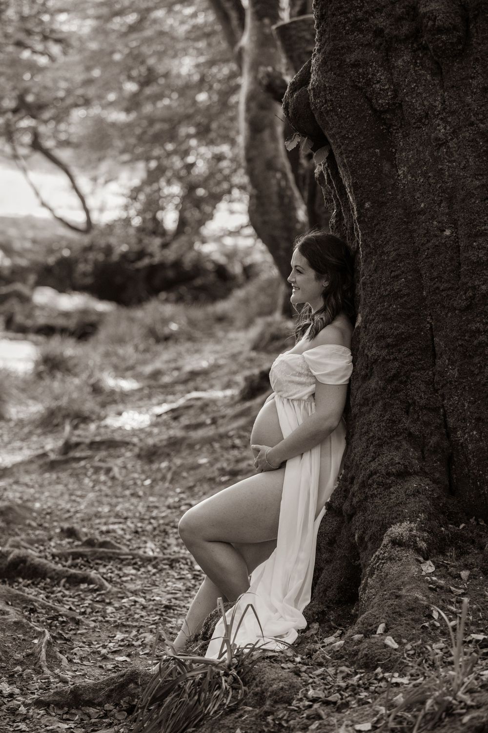 Black and white photo of pregnant woman in white dress leaning against large tree trunk in forest Cornwall Maternity Photographer