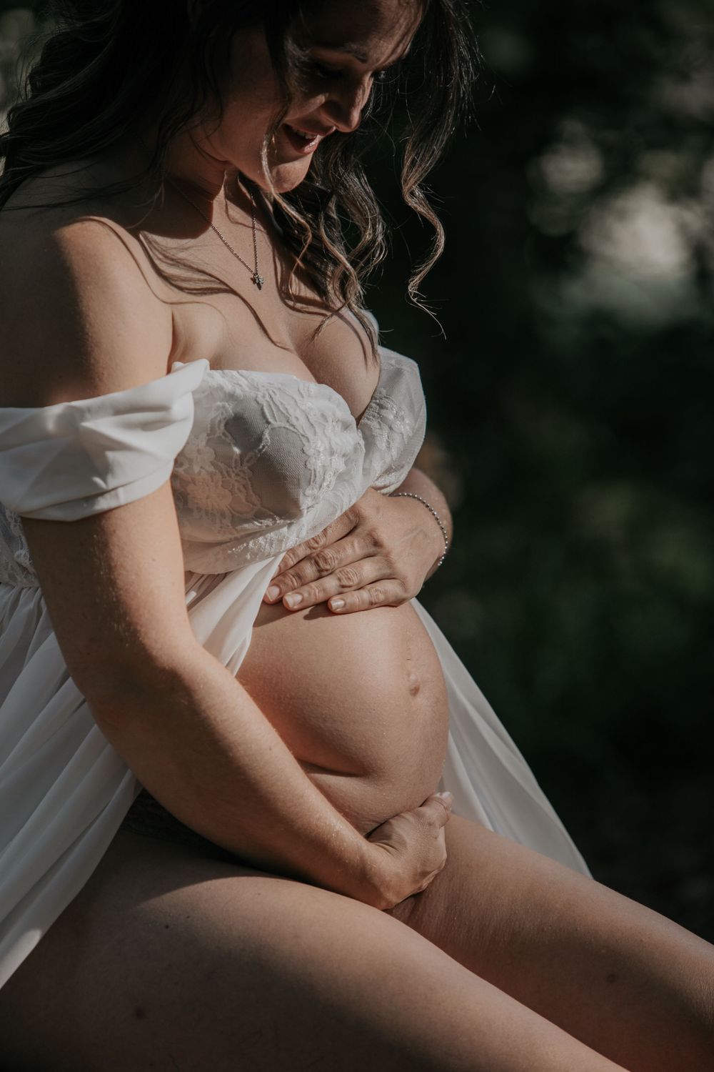 Pregnant woman in white off-shoulder lace gown smiling down while cradling her bare baby bump outdoors Cornwall Maternity Photographer
