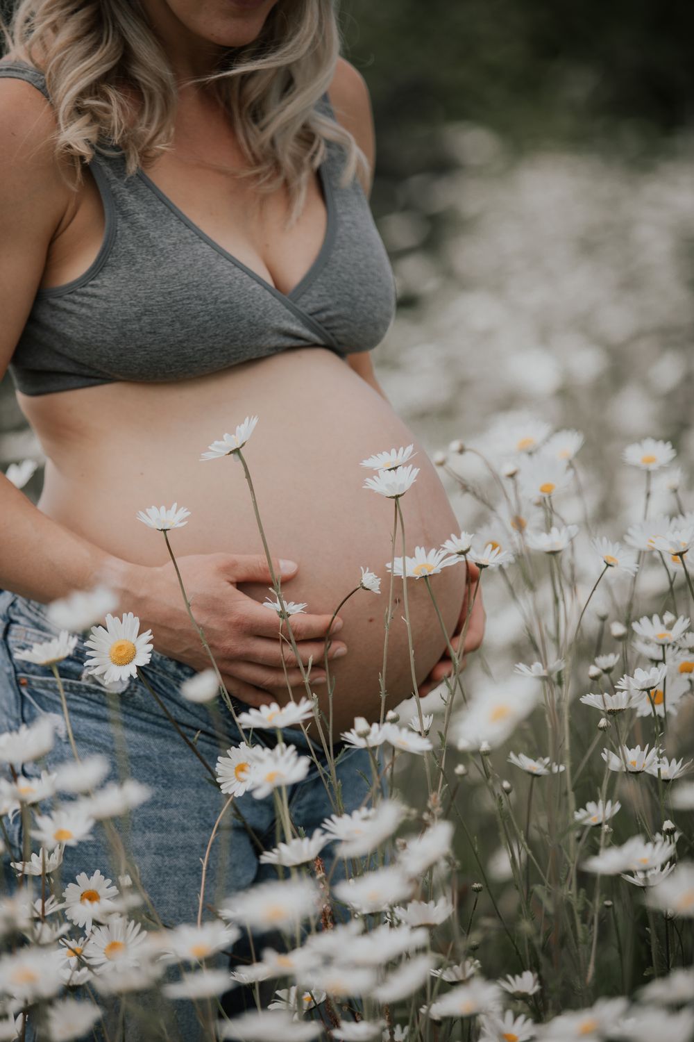 Pregnant woman in gray bralette and jeans holding her baby bump surrounded by white daisy wildflowers. Cornwall Maternity Photographer