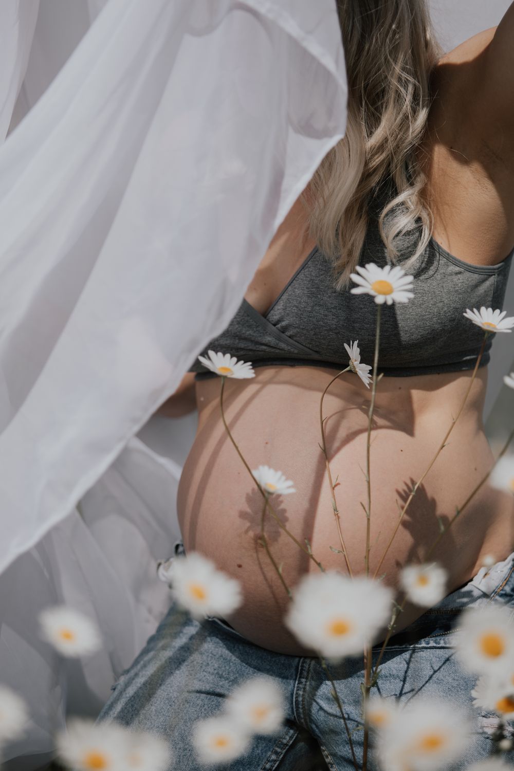 Pregnant woman in gray sports bra and jeans sitting among white daisy flowers in soft sunlight. Cornwall Maternity Photographer