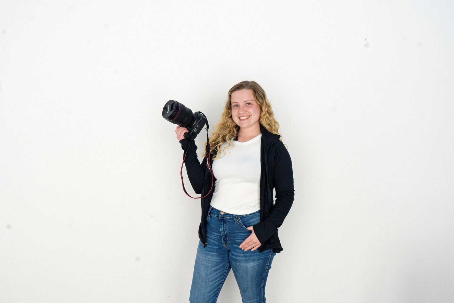 Smiling female photographer holding a DSLR camera, wearing a black cardigan and jeans, posing against a white wall.