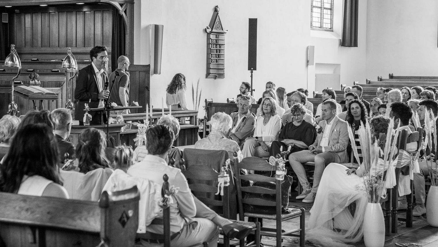 Zwart-witfoto van bruiloftsgasten die tijdens een ceremonie in een kerk zitten, met een spreker vooraan.