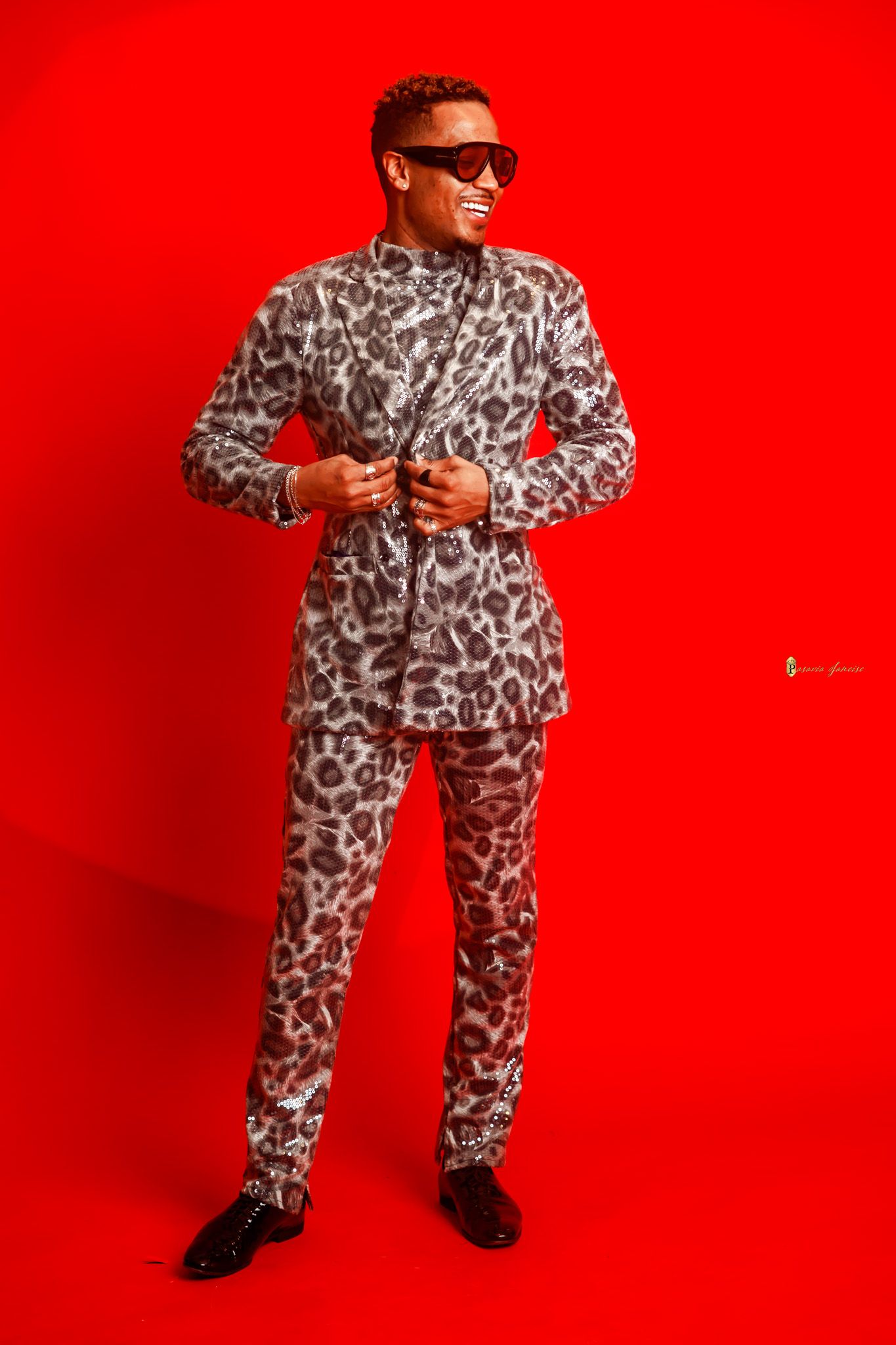 Man in leopard print suit and red sunglasses posing confidently against a bold red background.