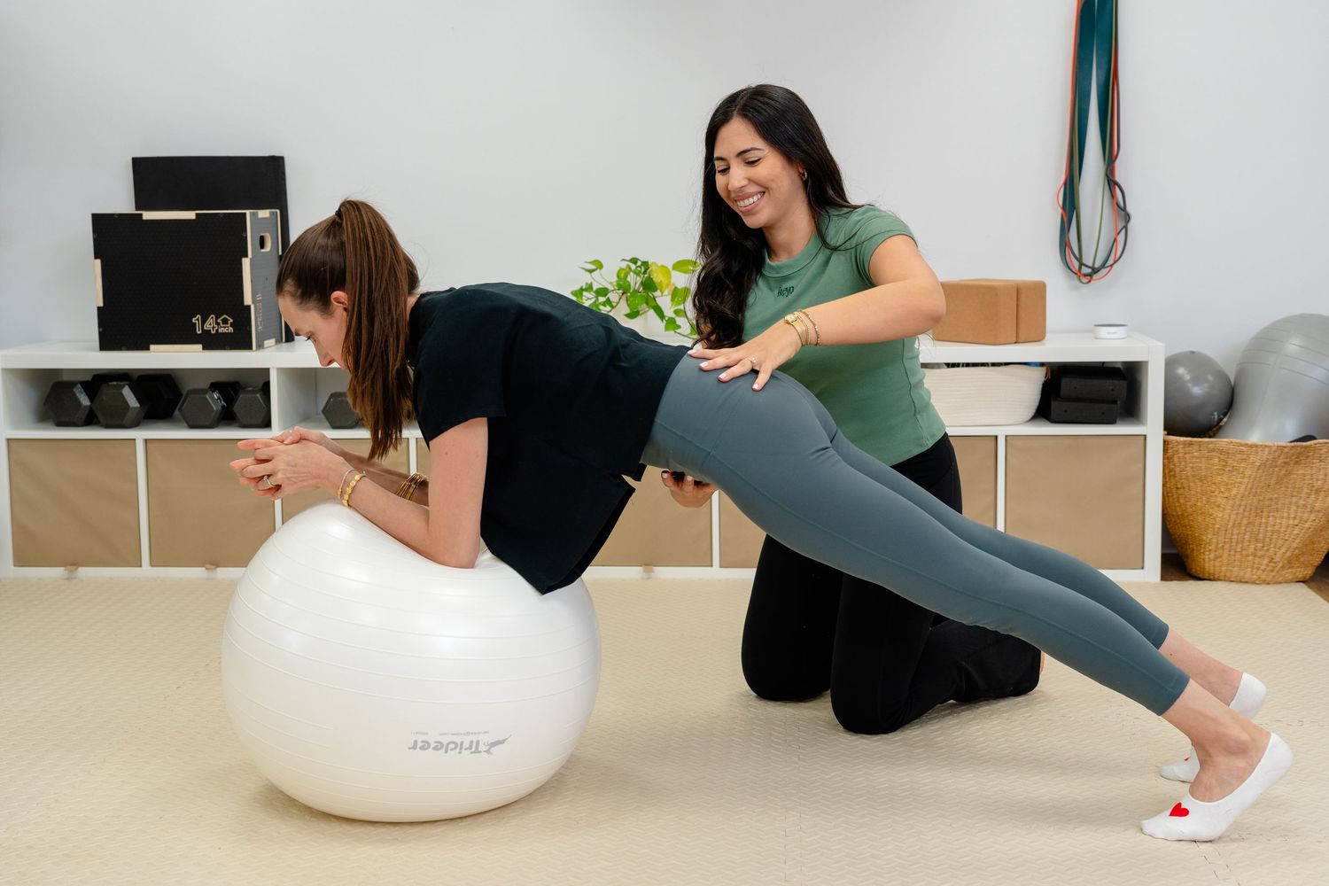 A trainer assists a woman doing a plank exercise on a white stability ball in a bright fitness studio.