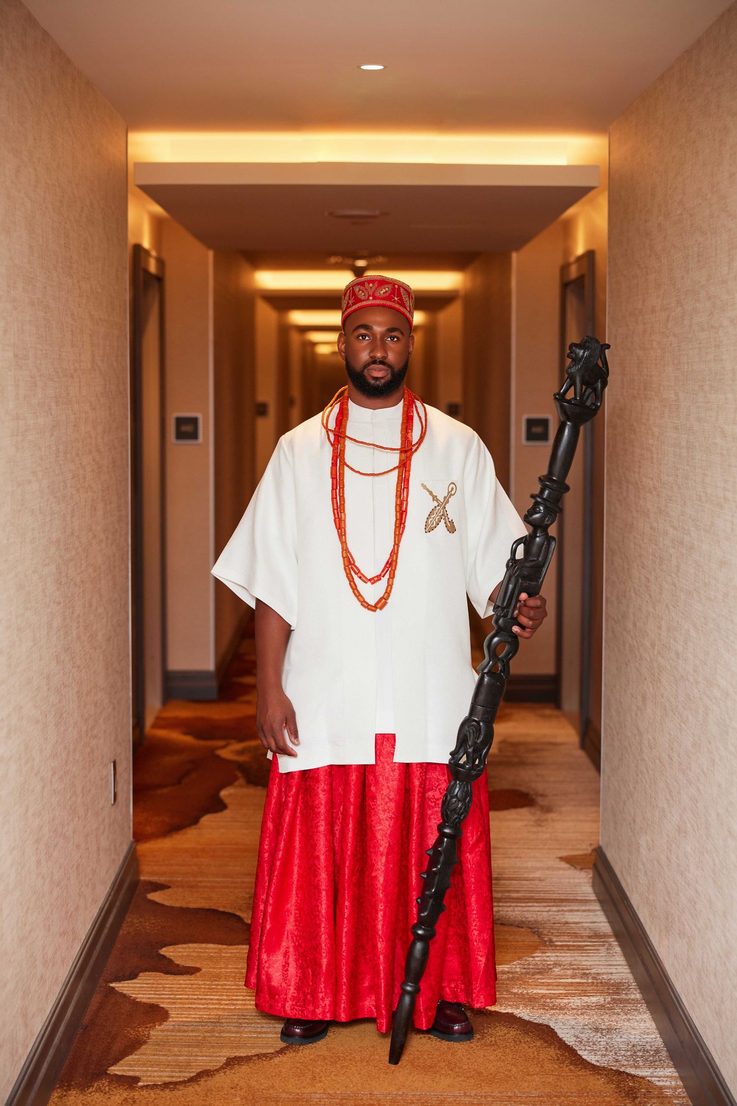 Man in traditional Nigerian attire with white top, red skirt, coral beads, and ceremonial staff in hotel hallway.