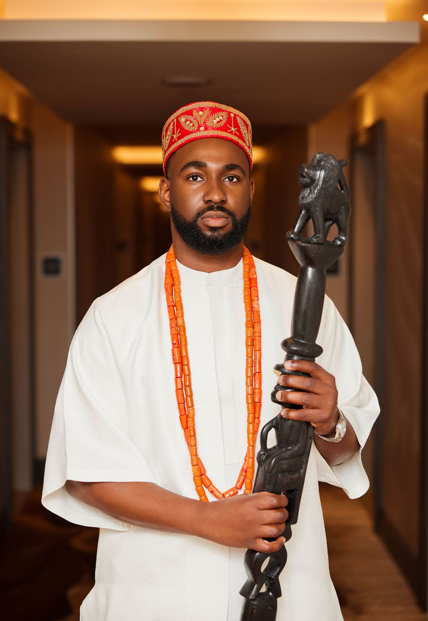Man in traditional Nigerian attire with coral beads, red cap, white agbada, holding carved wooden staff.