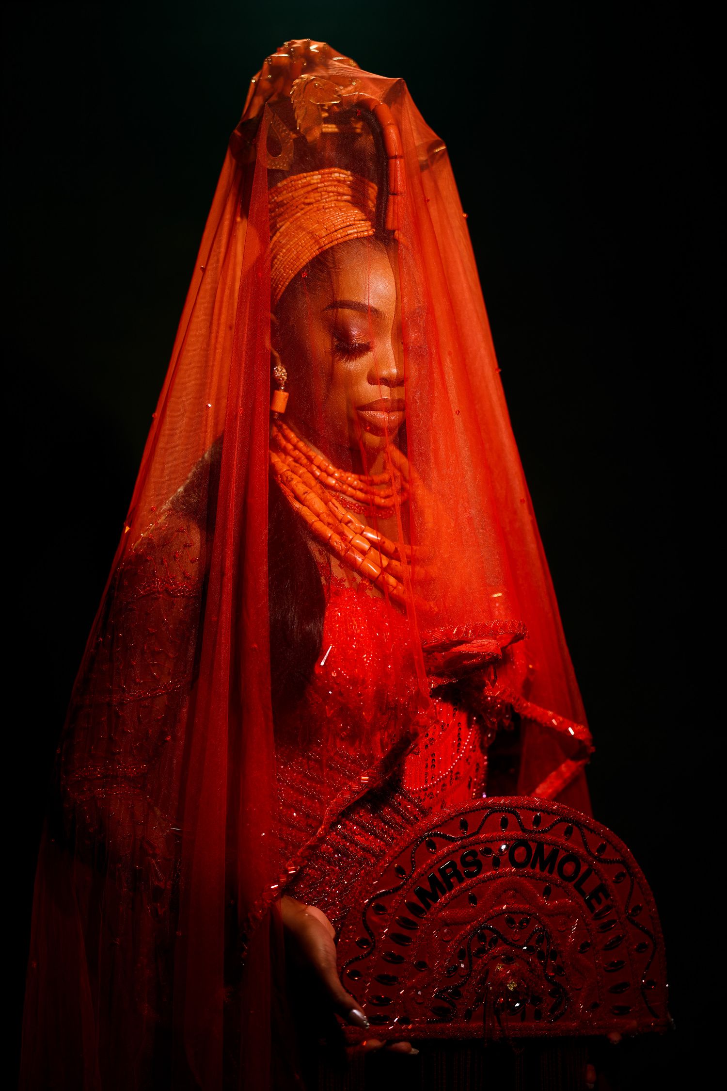 Woman in stunning red Nigerian bridal attire with gold headpiece, coral beads, and veil holding decorative fan.