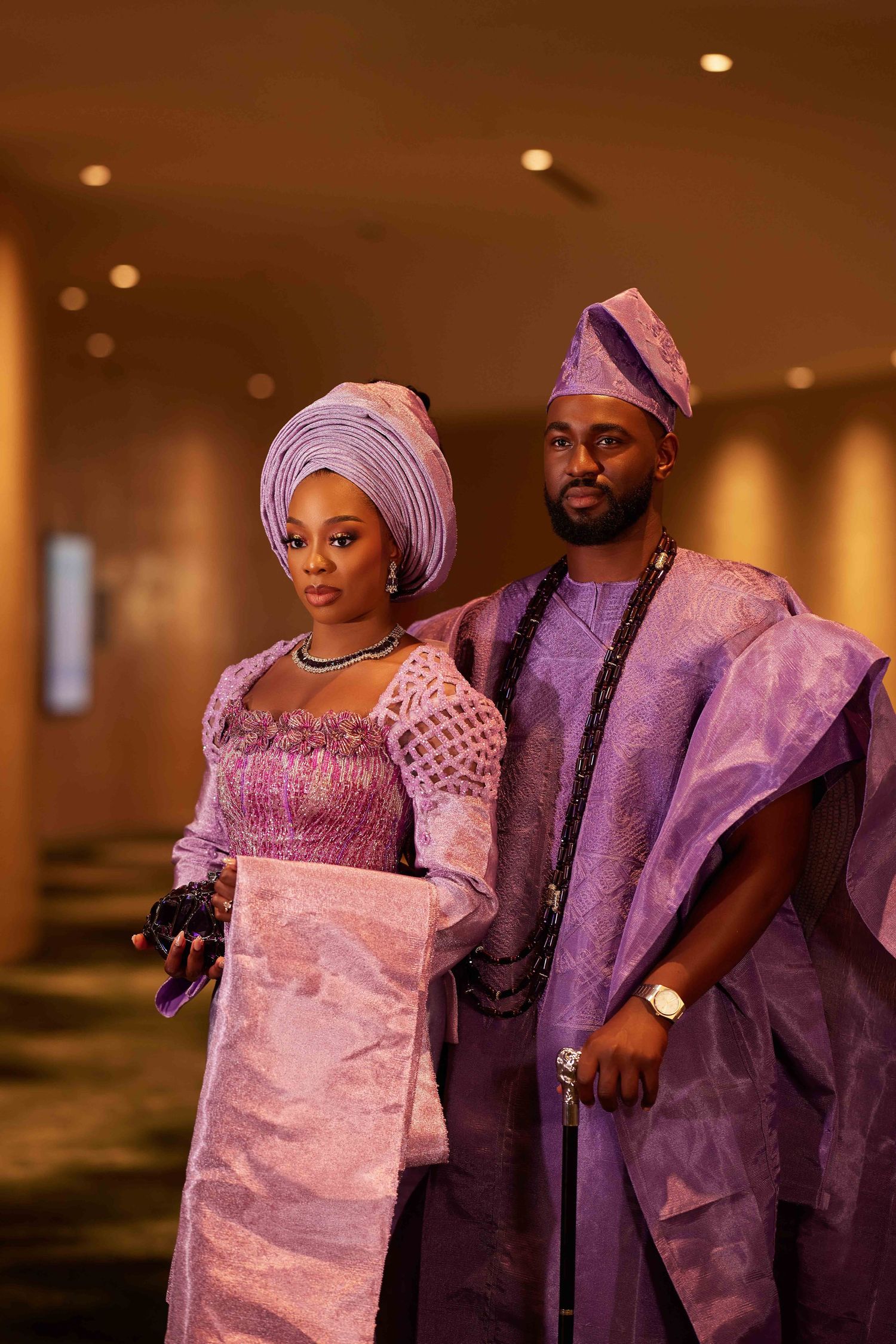 Couple in matching purple Nigerian traditional attire, woman in gele headwrap and man in agbada, posing indoors.