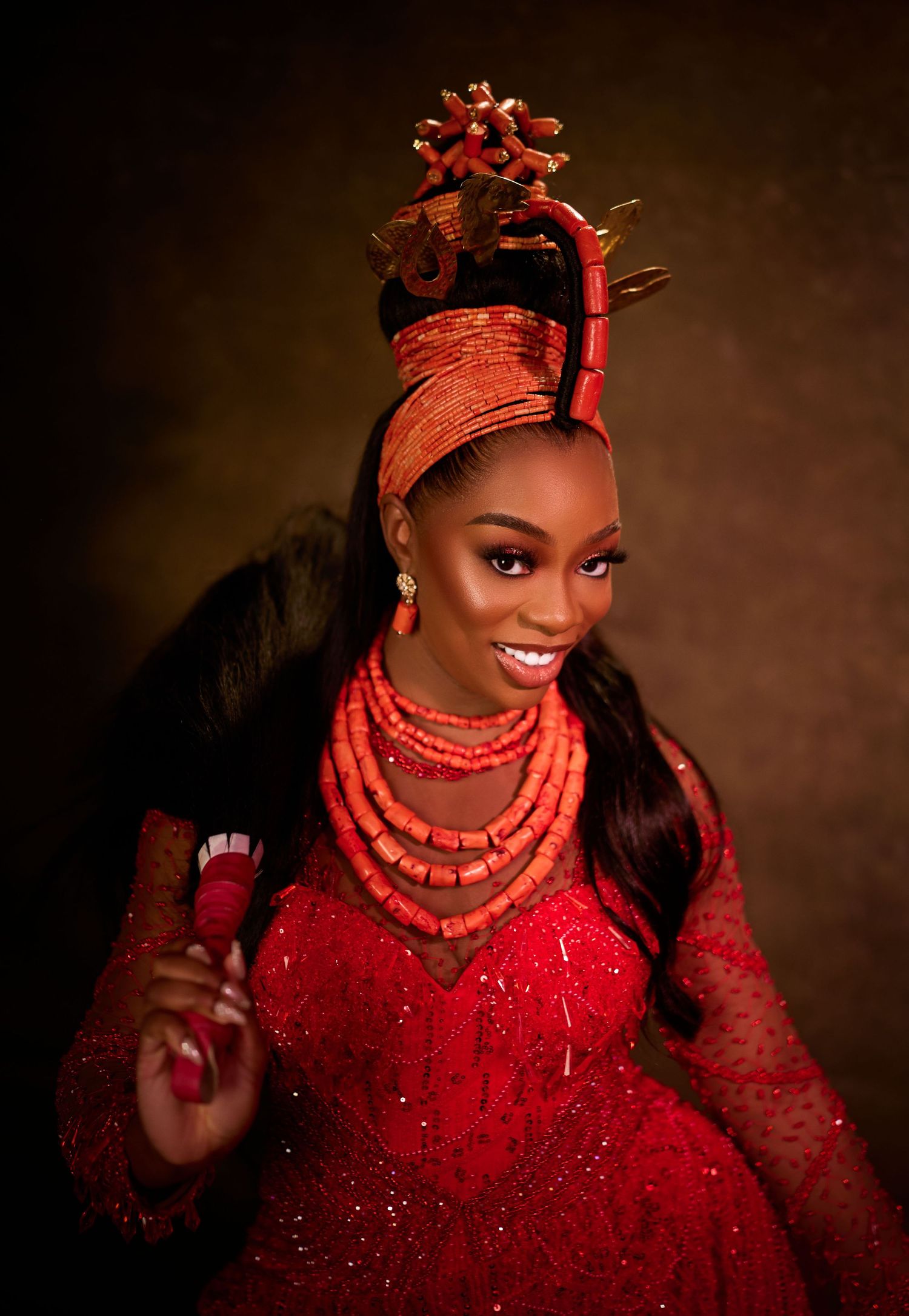 A woman in vibrant red Nigerian traditional bridal attire with coral beads and ornate headpiece smiling beautifully.