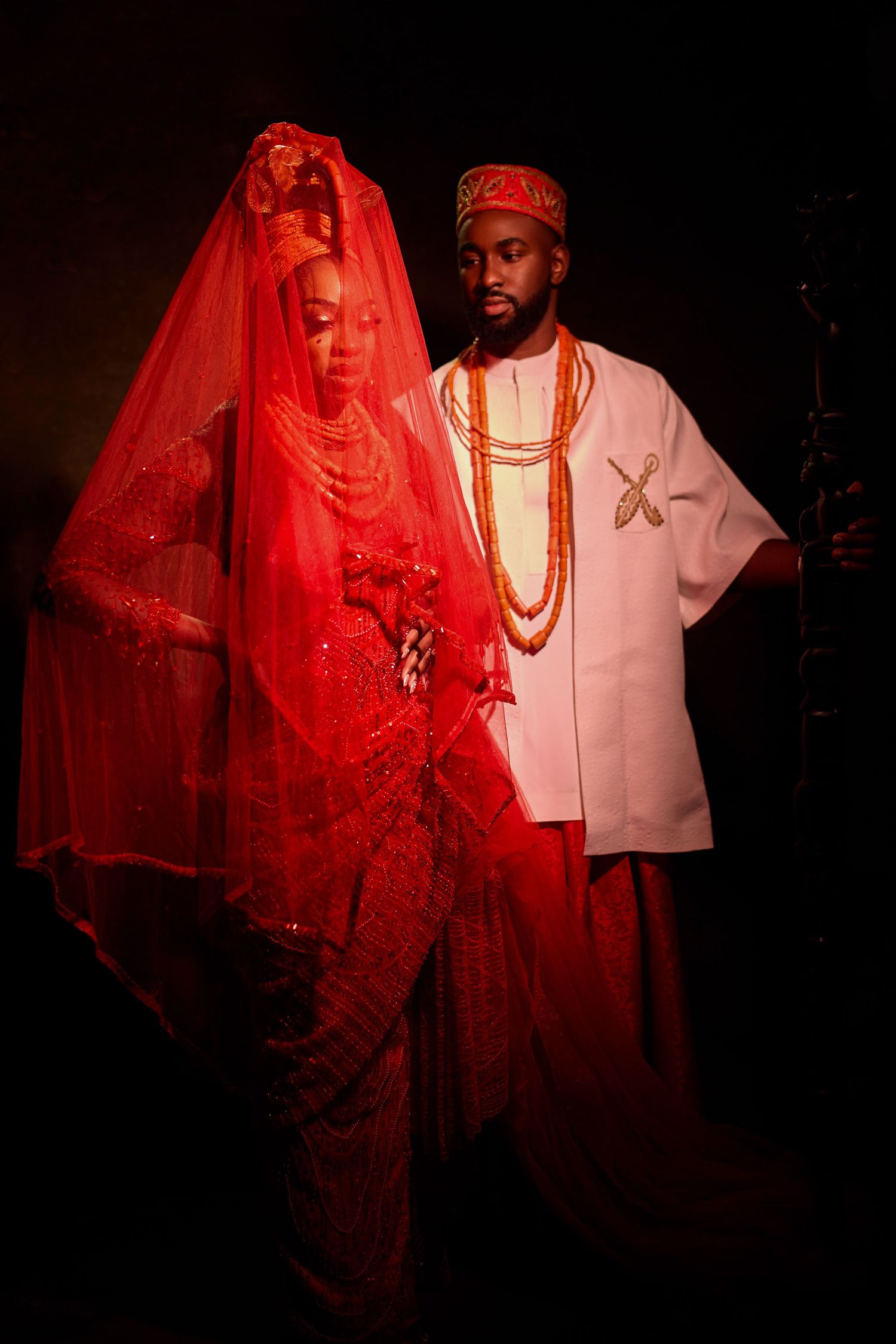 Couple in traditional Nigerian wedding attire, bride in red beaded gown with veil, groom in white with coral beads.