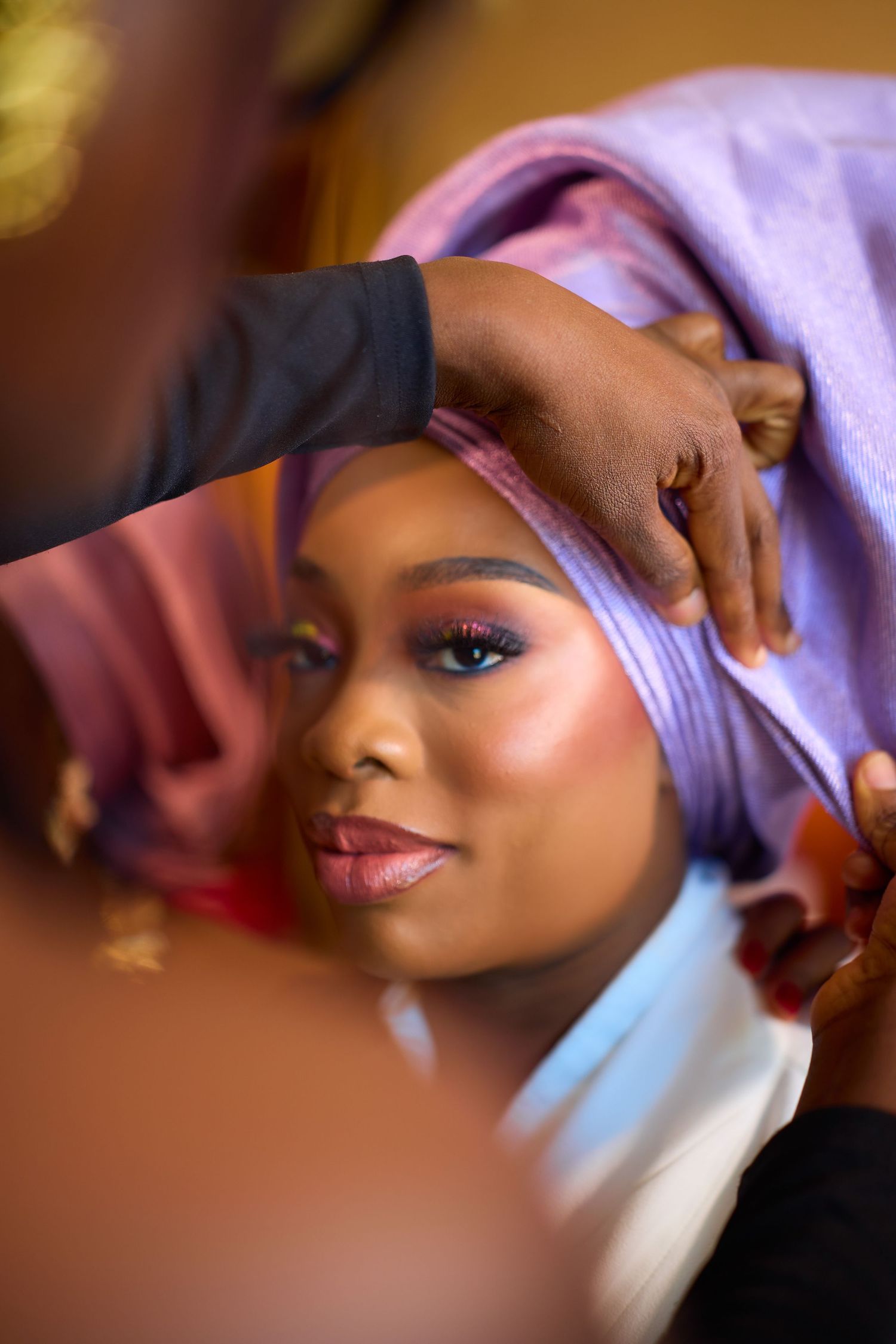 A makeup artist adjusting a purple head wrap on a woman with elegant eye makeup during bridal preparation.