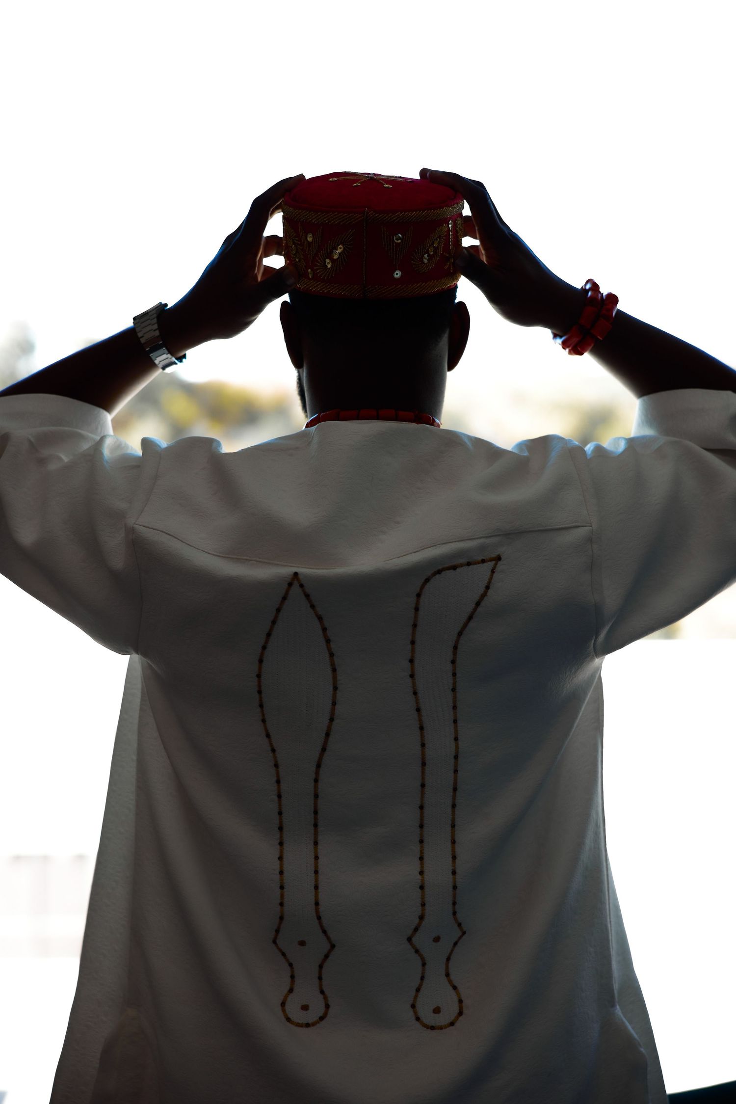 Man in white traditional robe placing a red kufi hat on his head, viewed from behind.