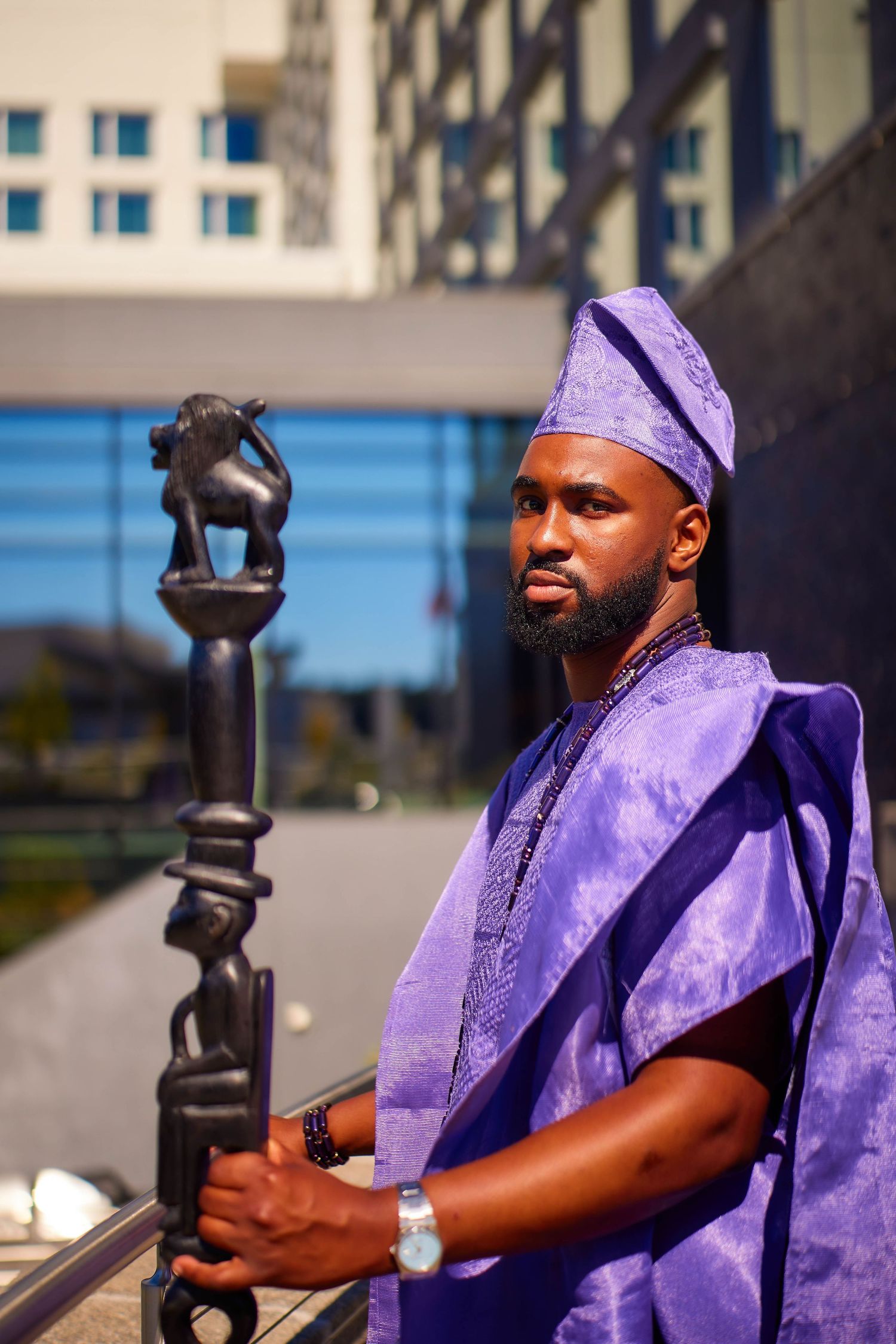 Man in purple traditional African attire holding carved staff poses confidently outdoors near modern building.