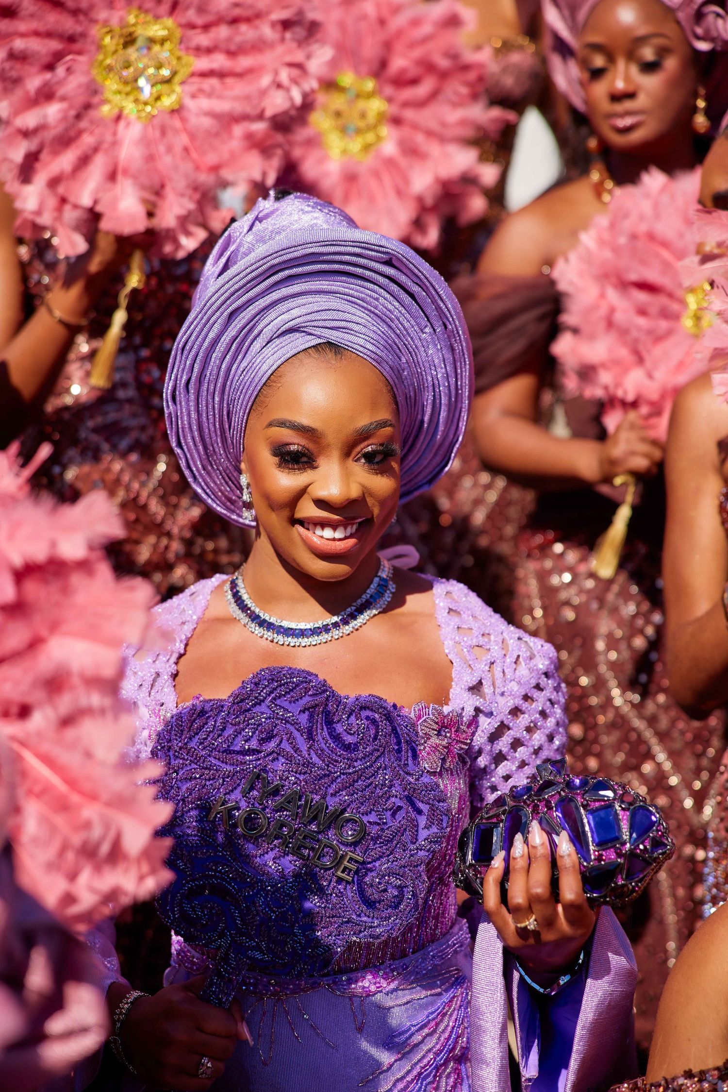 Smiling woman in purple Nigerian traditional attire with gele headwrap, holding clutch, surrounded by pink fans.
