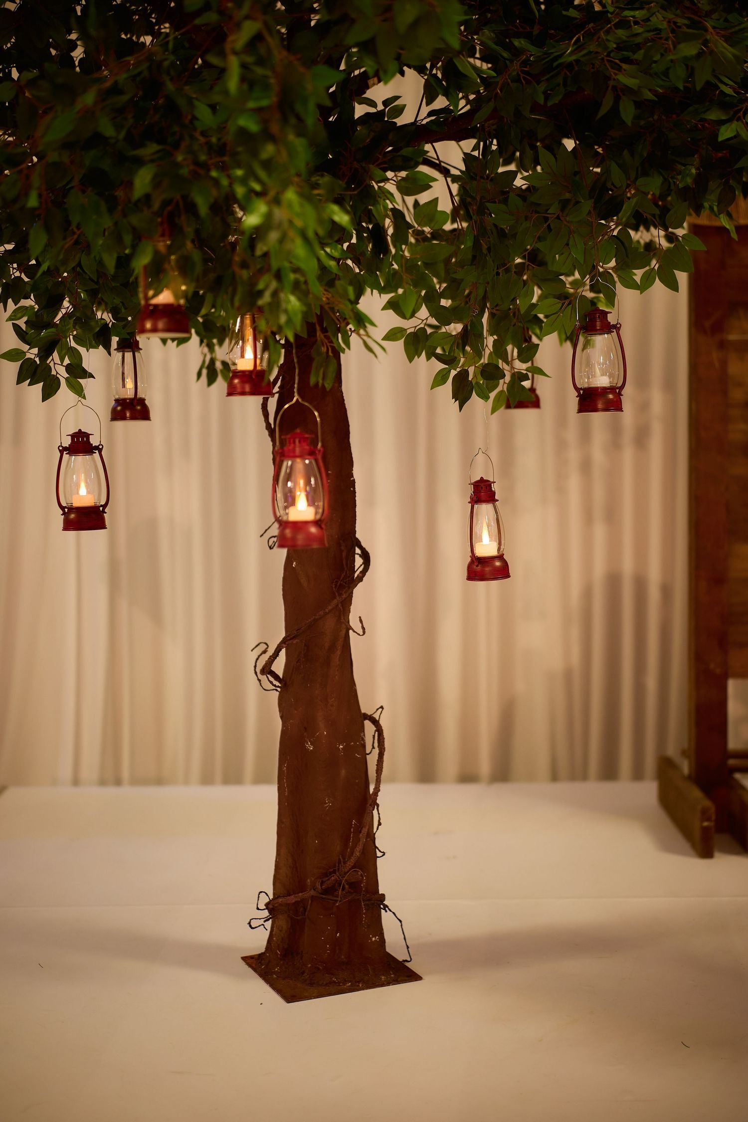 Decorative tree with hanging red lanterns glowing warmly, green leaves, and wrapped vines, set against a cream curtain backdrop.