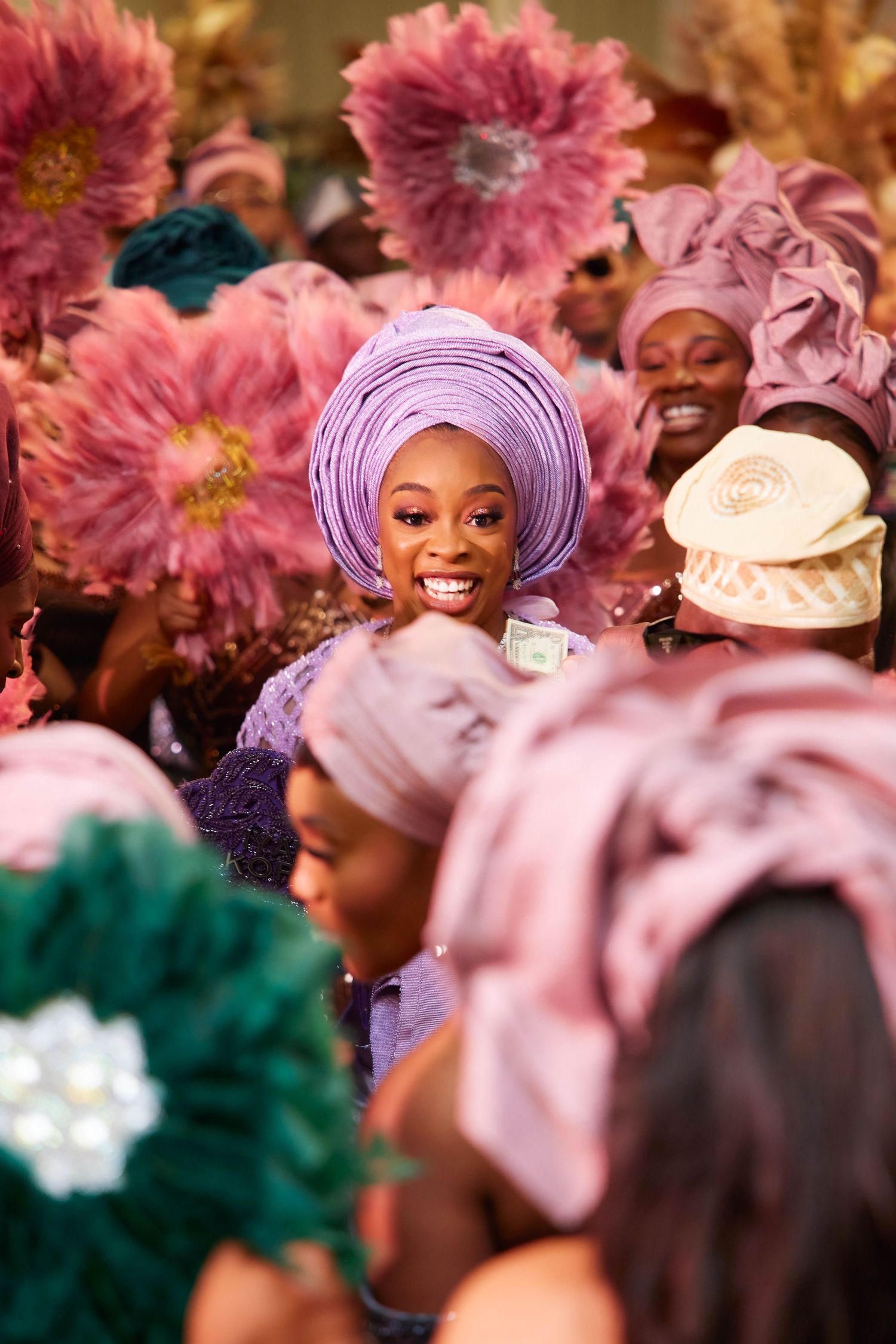 Smiling woman in lavender gele headwrap surrounded by women in pink traditional Nigerian attire at a festive celebration.