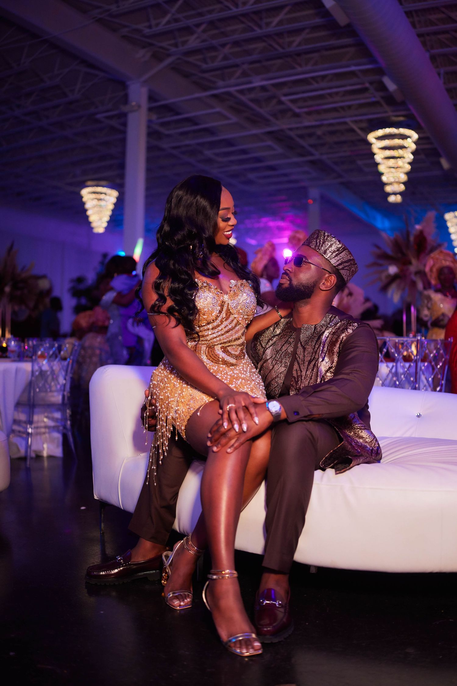 Elegant couple in formal attire sharing a romantic moment on a white sofa at a glamorous event venue.