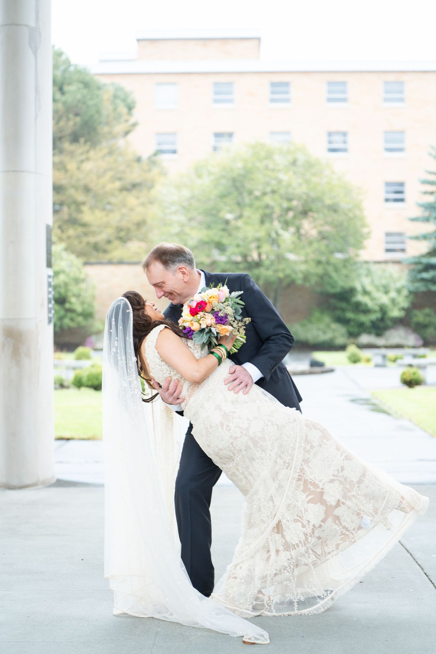 Bride and groom share a romantic dip kiss outdoors, bride in lace gown holding colorful bouquet, groom in black tuxedo.