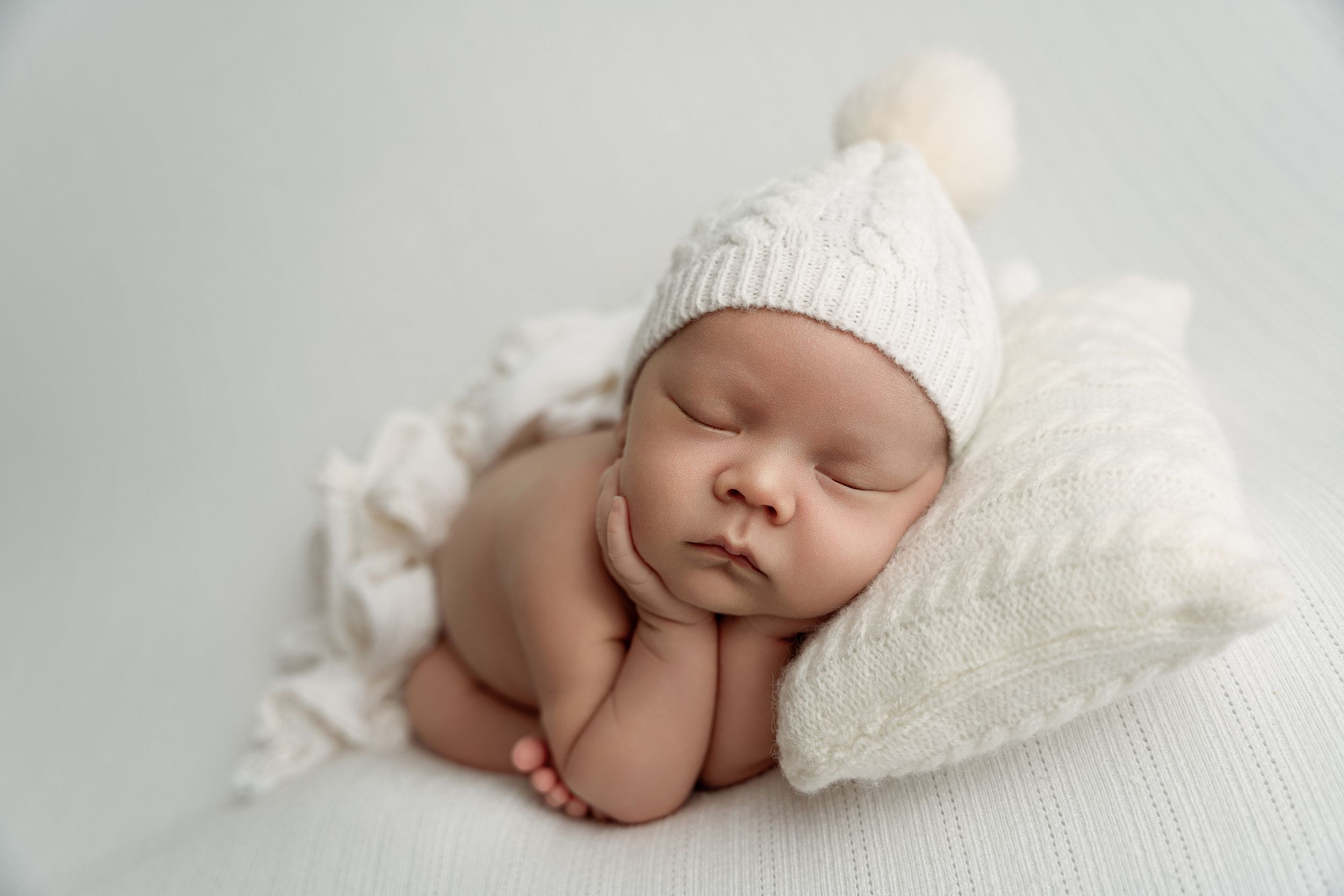 Peaceful newborn baby sleeping in white knit hat with pom-pom, resting on soft white blanket and pillow.