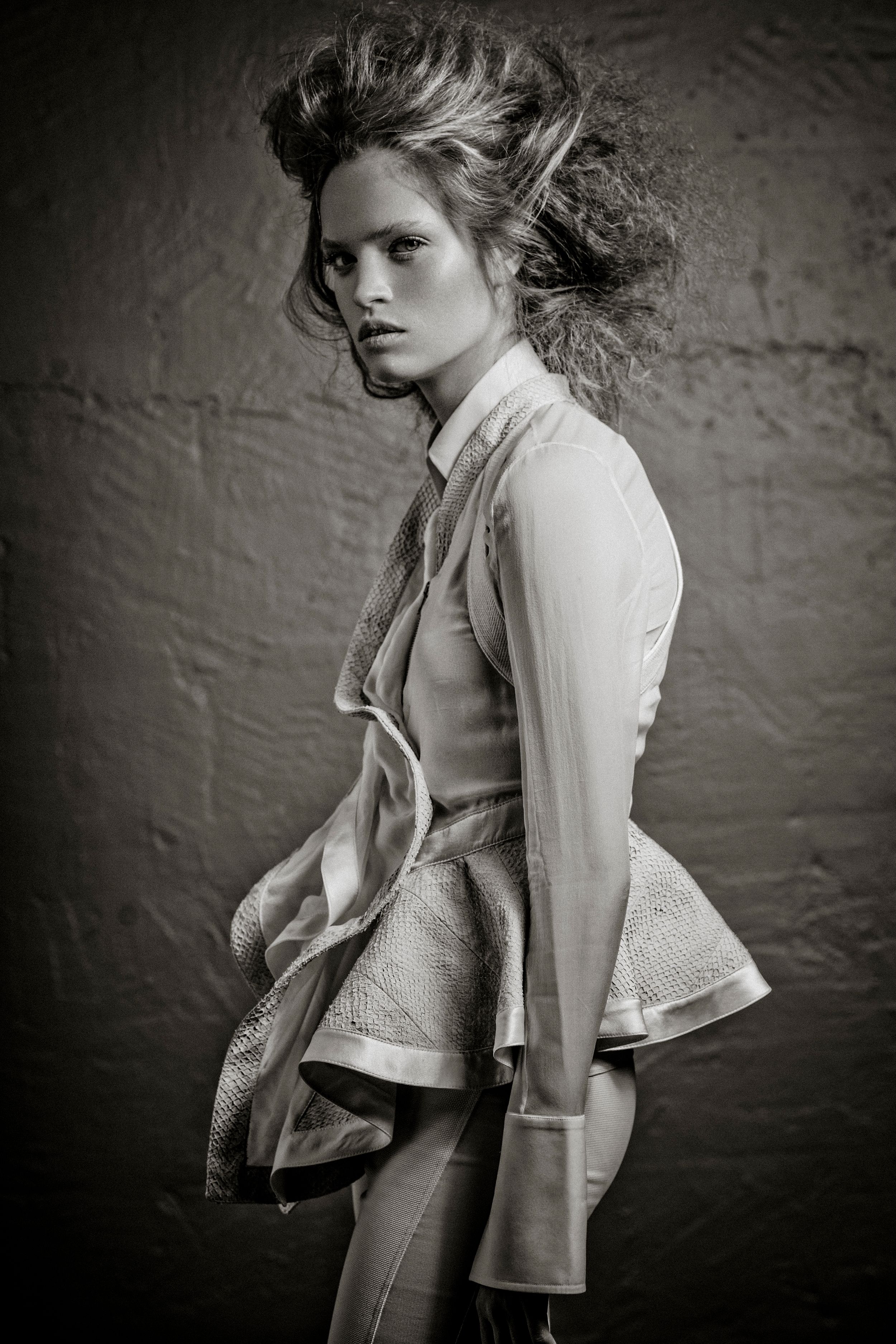 Black and white fashion photo of a young woman in stylish layered outfit with windswept hair posing against a textured wall.