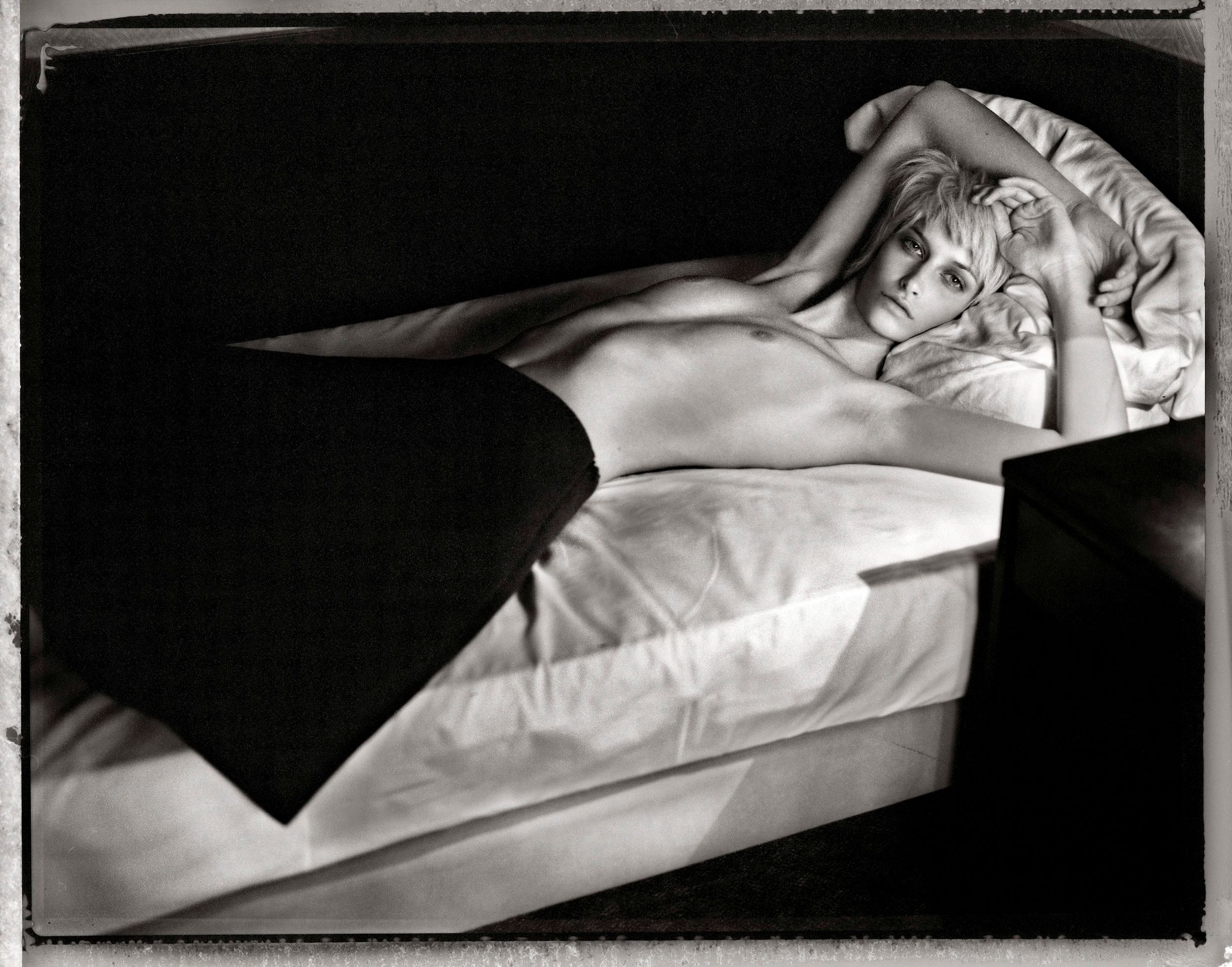 Black and white photo of a woman reclining on a bed, illuminated by dramatic light and shadow.
