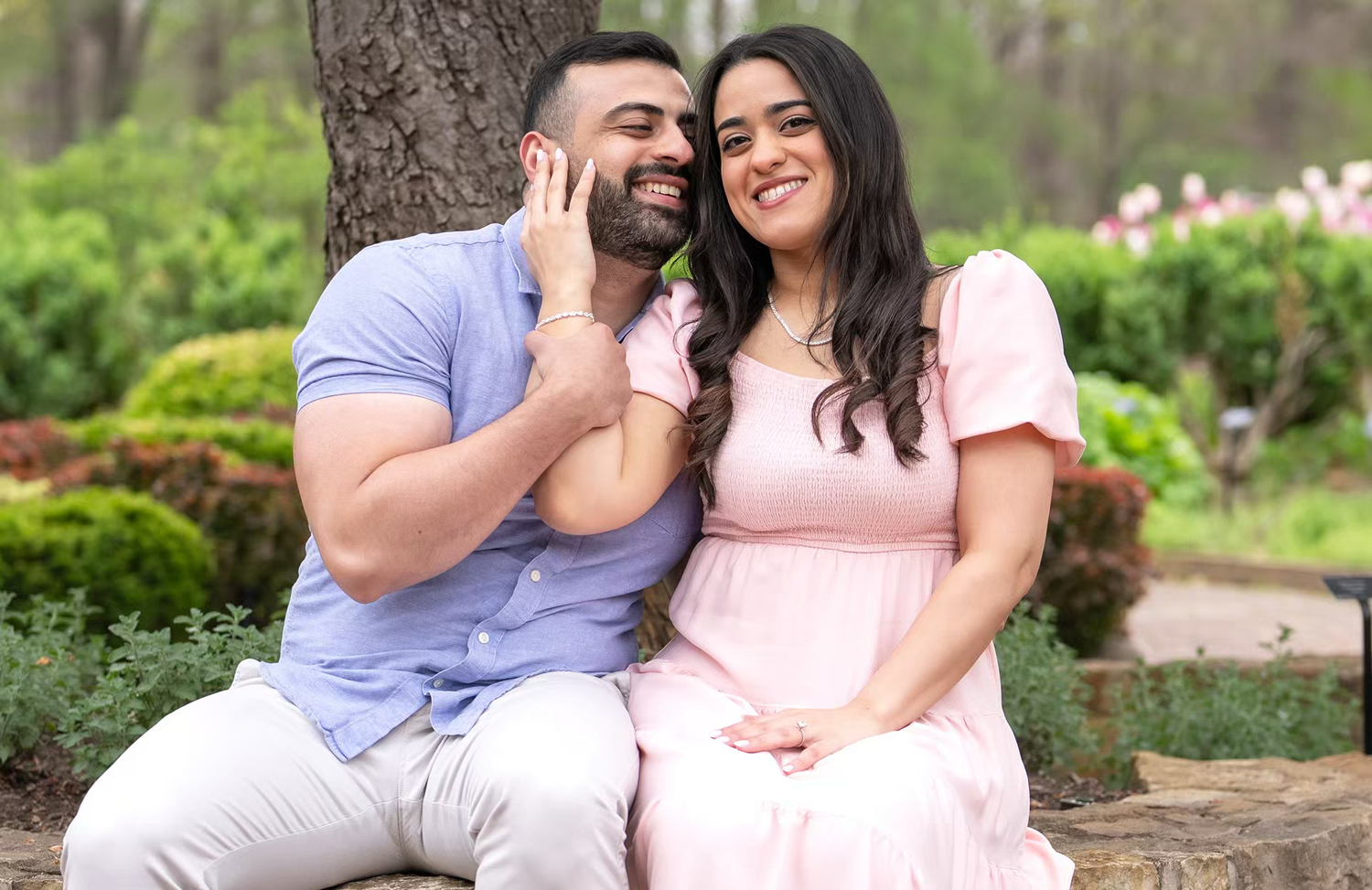 An engagement portrait of smiling couple sits outdoors near a tree, at Inniswood Metro Gardens