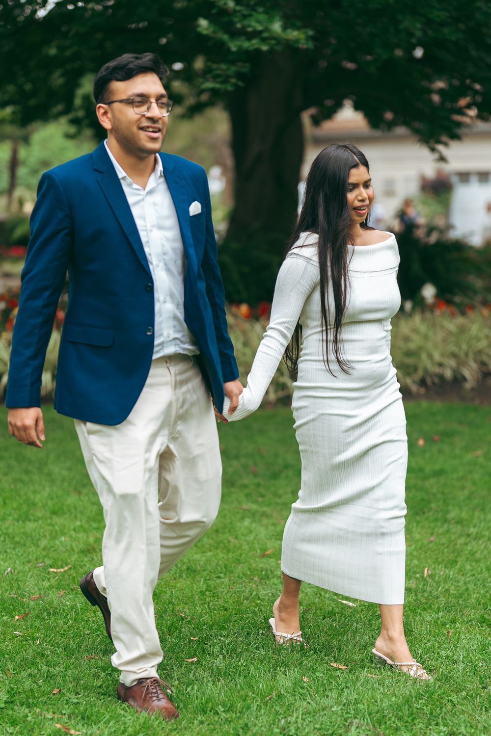 A couple walks hand-in-hand on green grass, man in blue blazer, woman in white midi dress, in a garden setting.