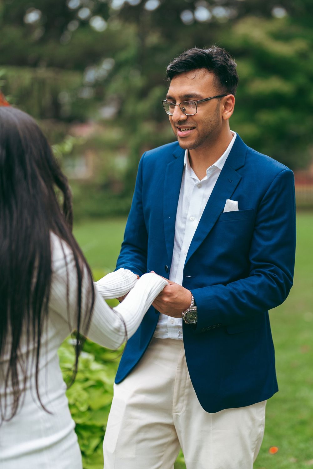 Young man in blue blazer and glasses smiling while exchanging items outdoors in a garden setting.