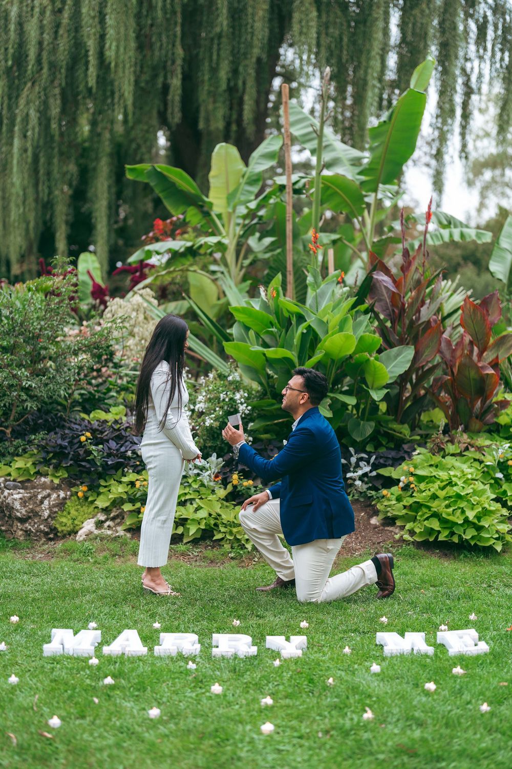 Man proposes on one knee in lush garden with Marry Me spelled out on grass while woman stands before him.