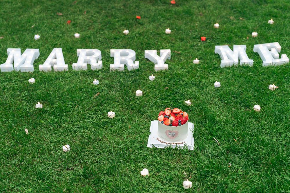 Romantic marriage proposal setup with white MARRY ME letters and a strawberry cake on lush green grass.