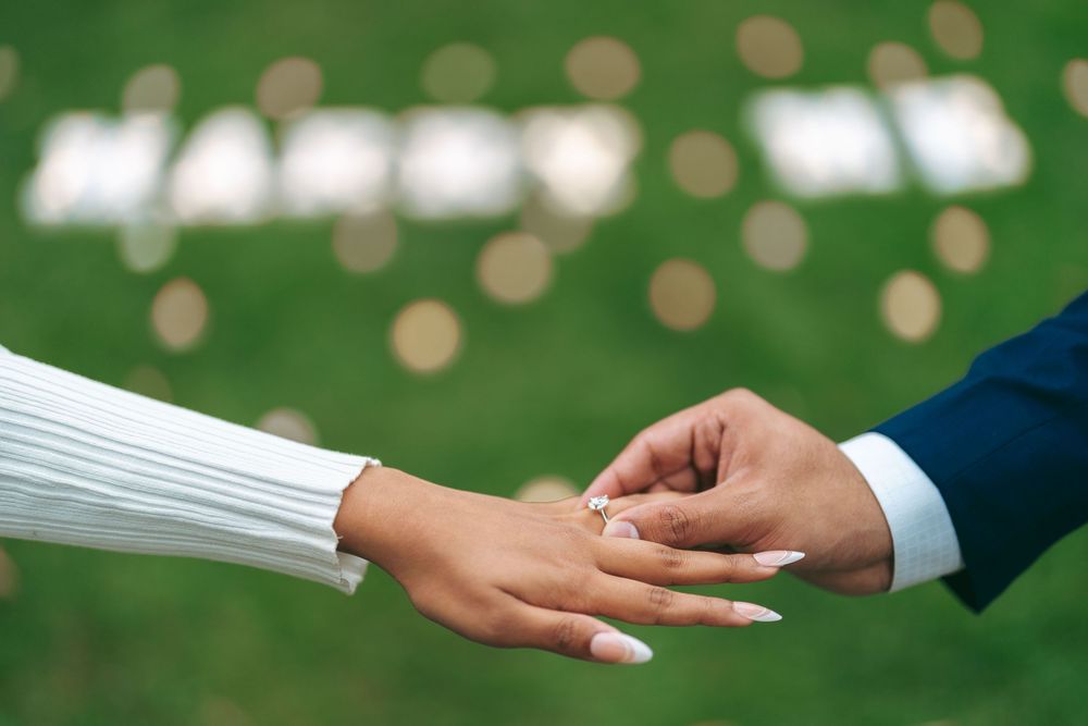 A groom placing a diamond engagement ring on his bride's hand outdoors with a Marry Me sign in the background.