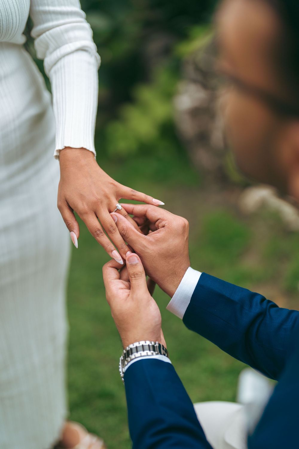 Groom in blue suit placing wedding ring on bride's finger outdoors during ceremony.