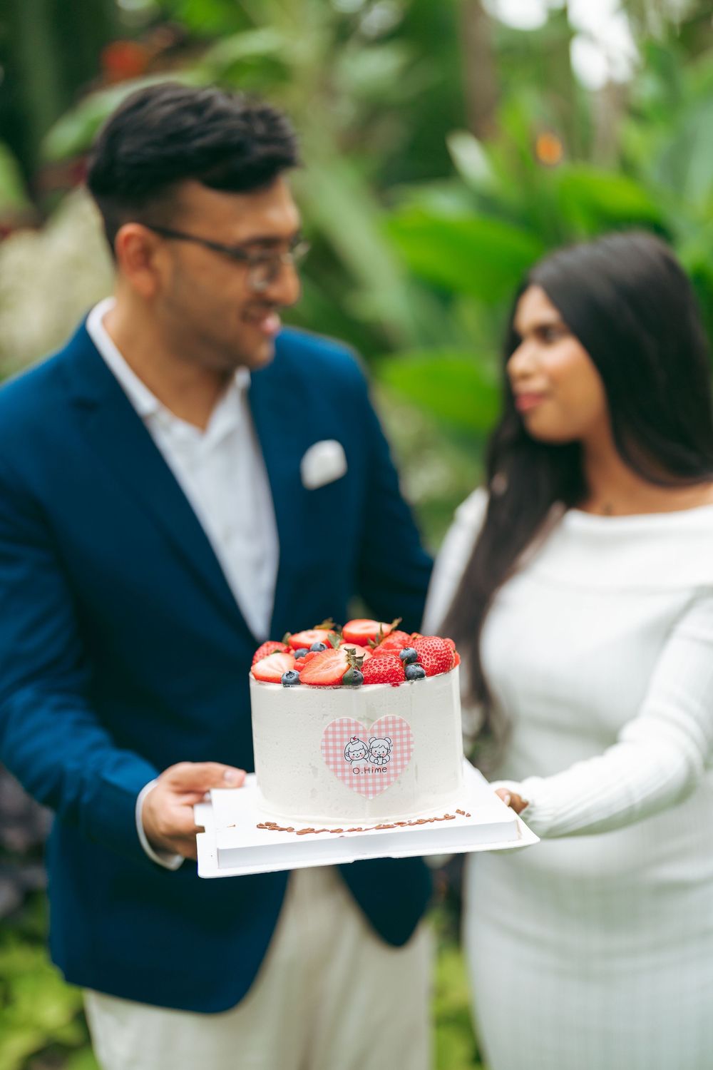 Couple holding a white cake topped with fresh strawberries and berries outdoors in a lush garden setting.