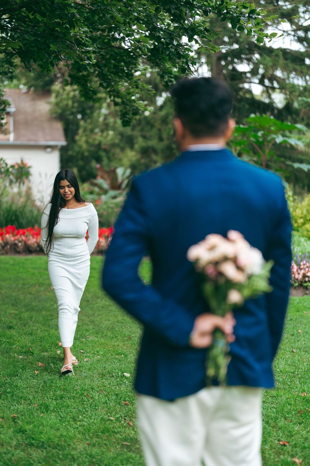 Man in blue suit hiding flowers behind his back for woman in white dress walking toward him in garden.