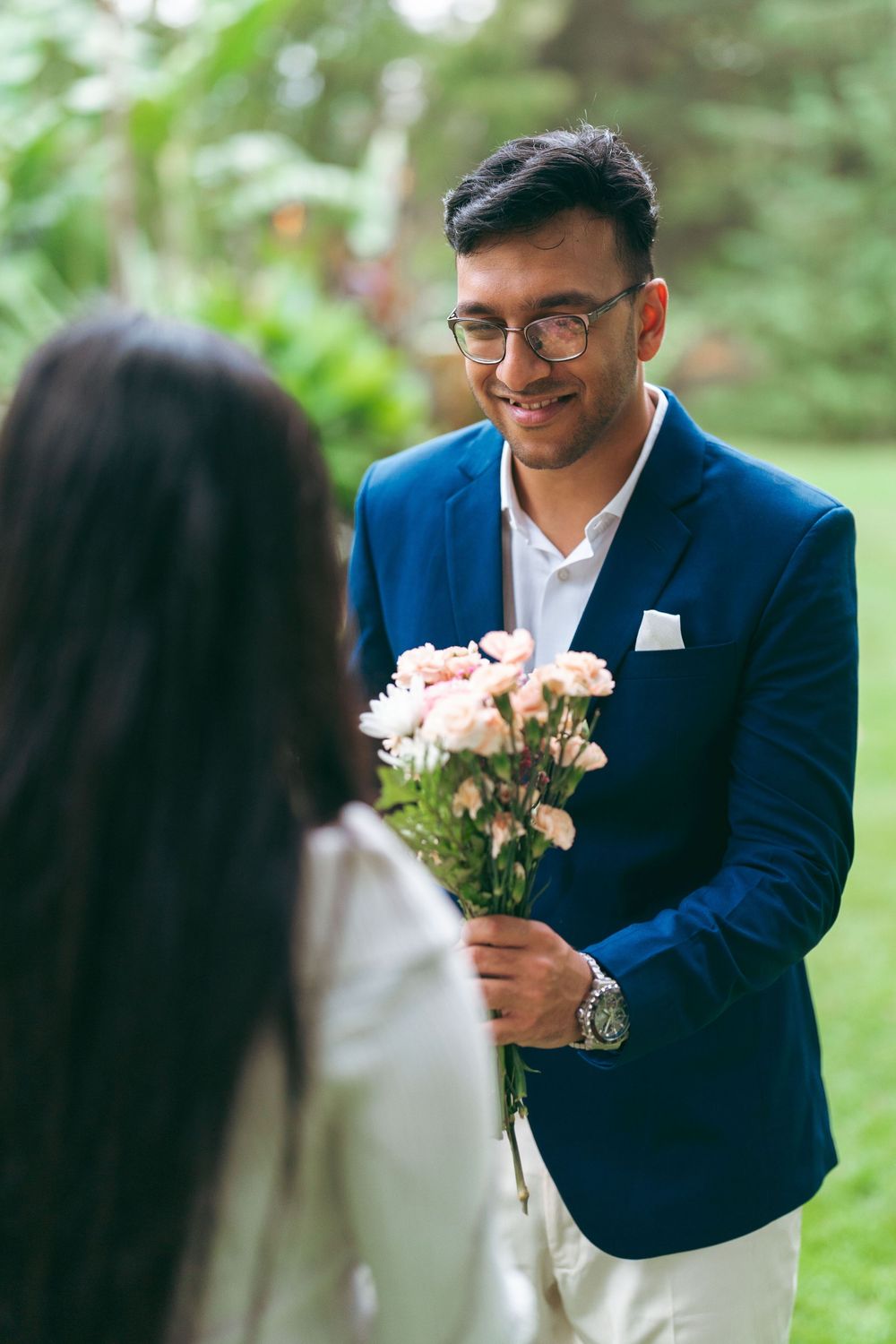 Smiling man in blue blazer presenting pink roses bouquet to woman outdoors in garden setting.