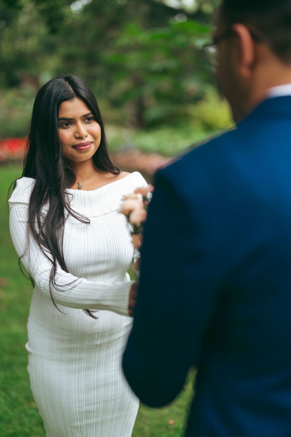 Woman in white ribbed dress holds hands with man in blue suit outdoors in green garden setting.