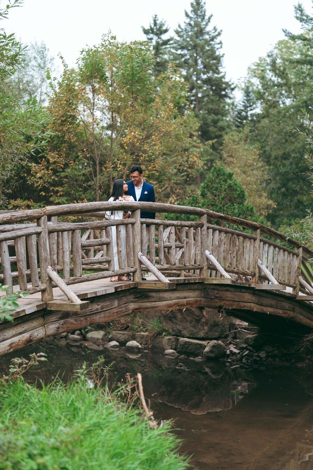 Couple standing on a rustic wooden bridge over a stream surrounded by lush green trees.