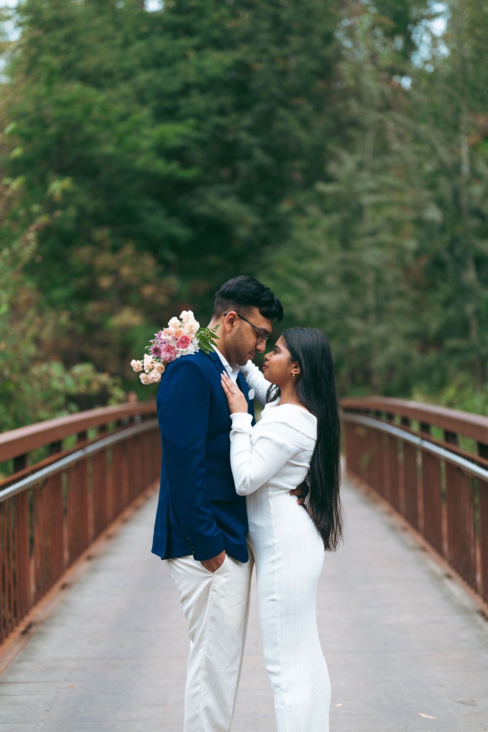 Couple in wedding attire embracing on a red bridge surrounded by lush green trees, holding a pink floral bouquet.