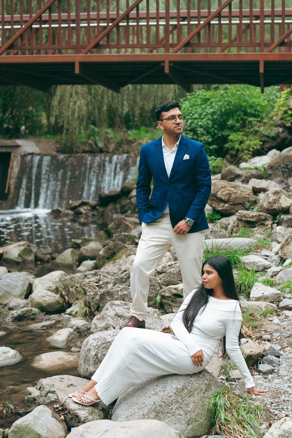 Couple posing on rocks near waterfall, man in blue blazer standing, woman in white dress sitting, lush green background.