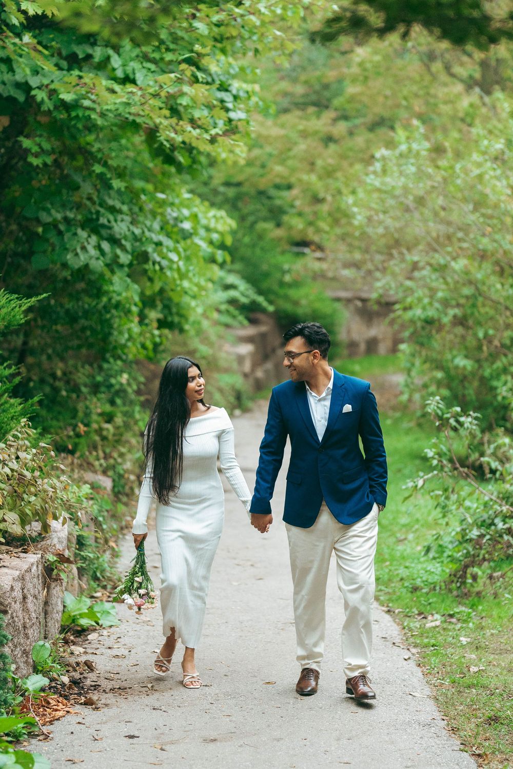Couple holding hands walking on a garden path, woman in white dress and man in blue blazer with white pants.