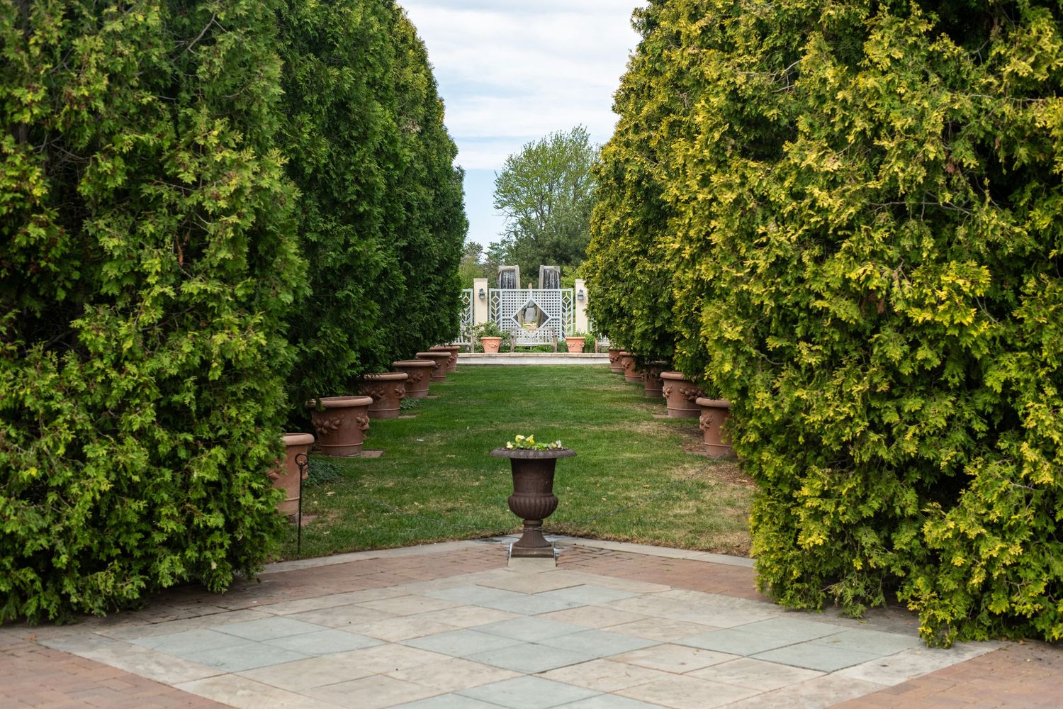 Formal garden pathway flanked by tall green hedges leading to a white ornamental structure with potted urns on a brick terrace.