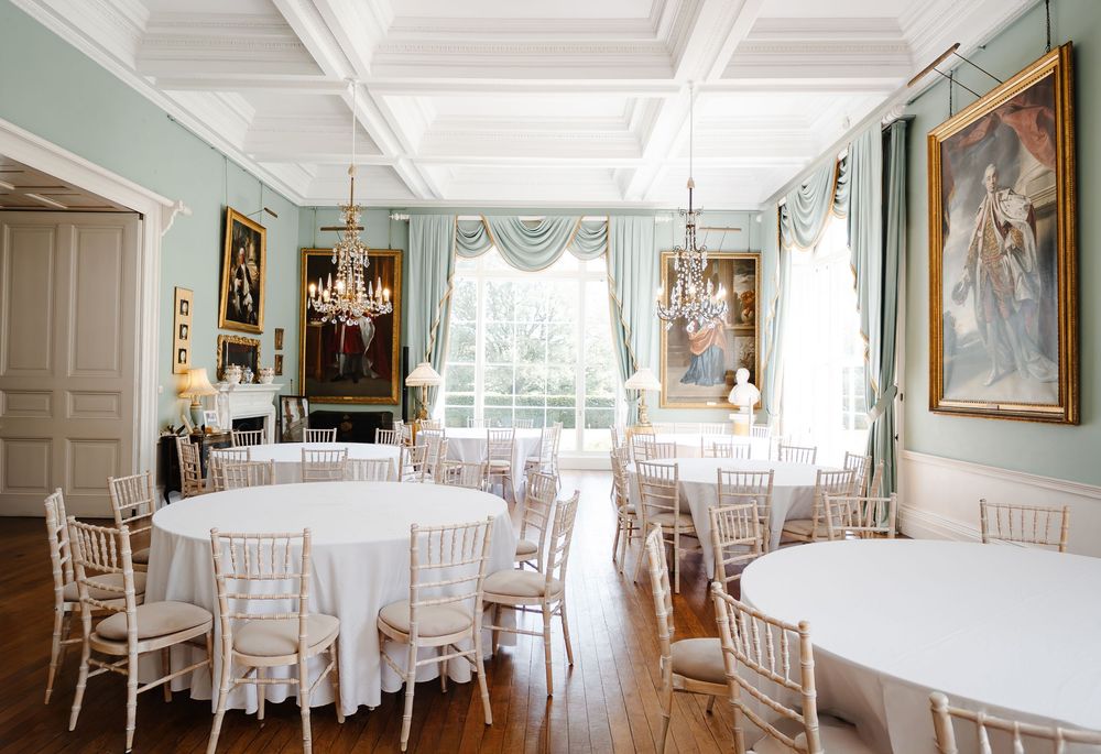 Elegant Georgian dining room with round tables, chiavari chairs, chandeliers, and pastel blue walls with portrait paintings.