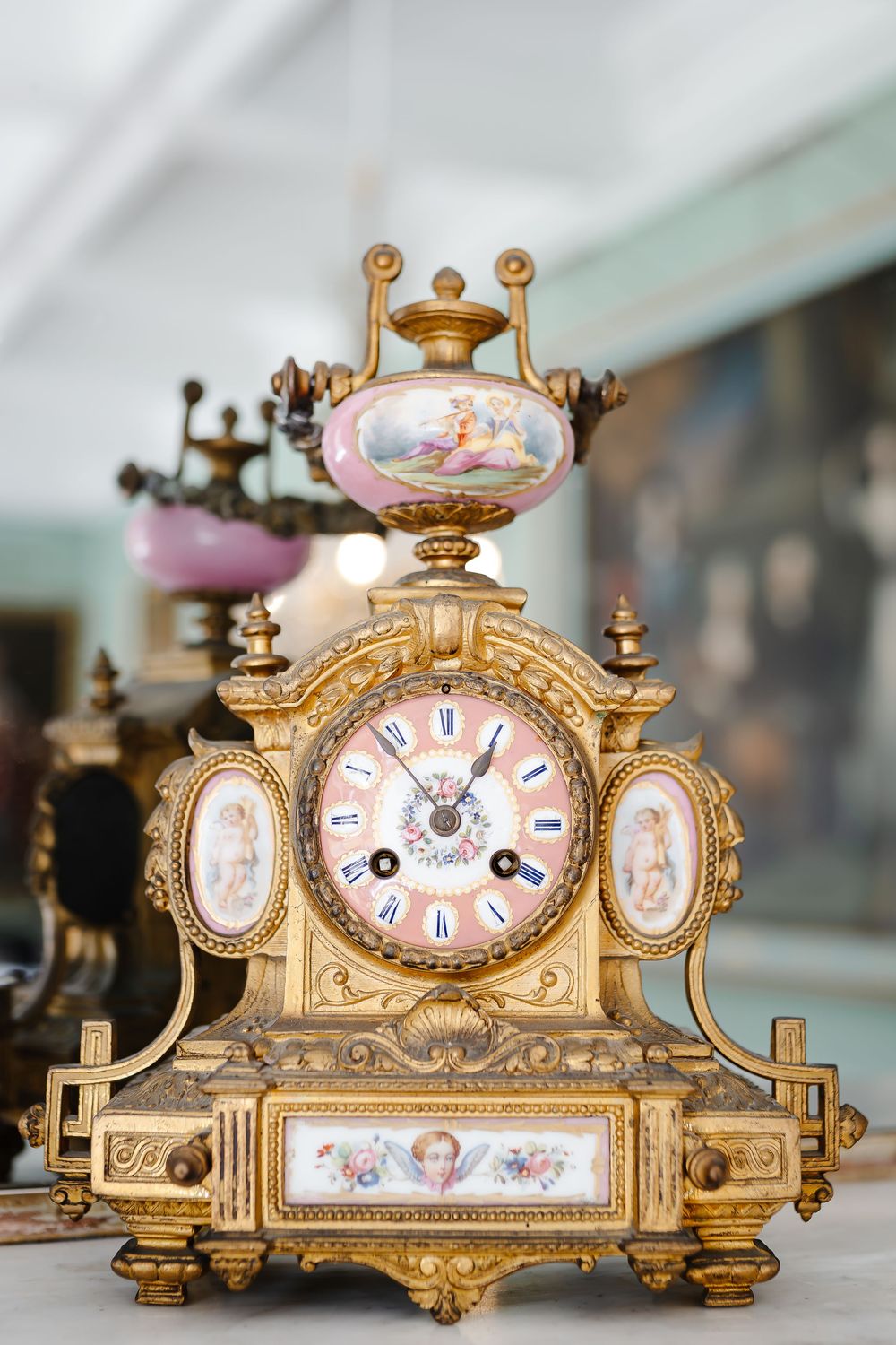 Ornate gilt French mantel clock with porcelain panels featuring cherub paintings and Roman numeral dial.