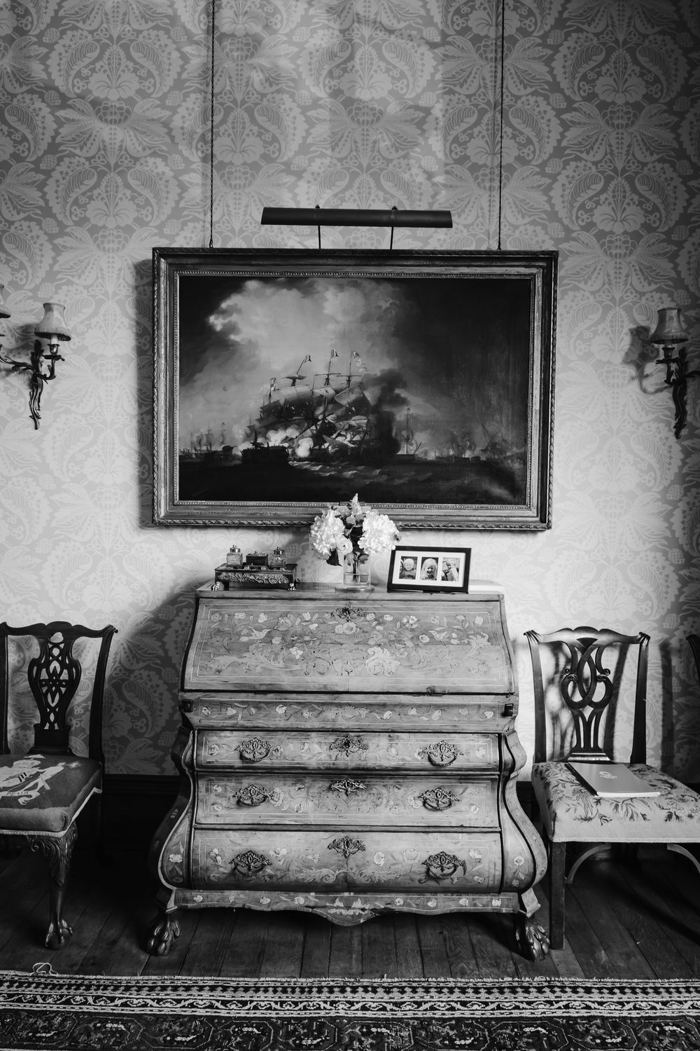Black and white photo of antique bureau with naval battle painting above flanked by chairs.