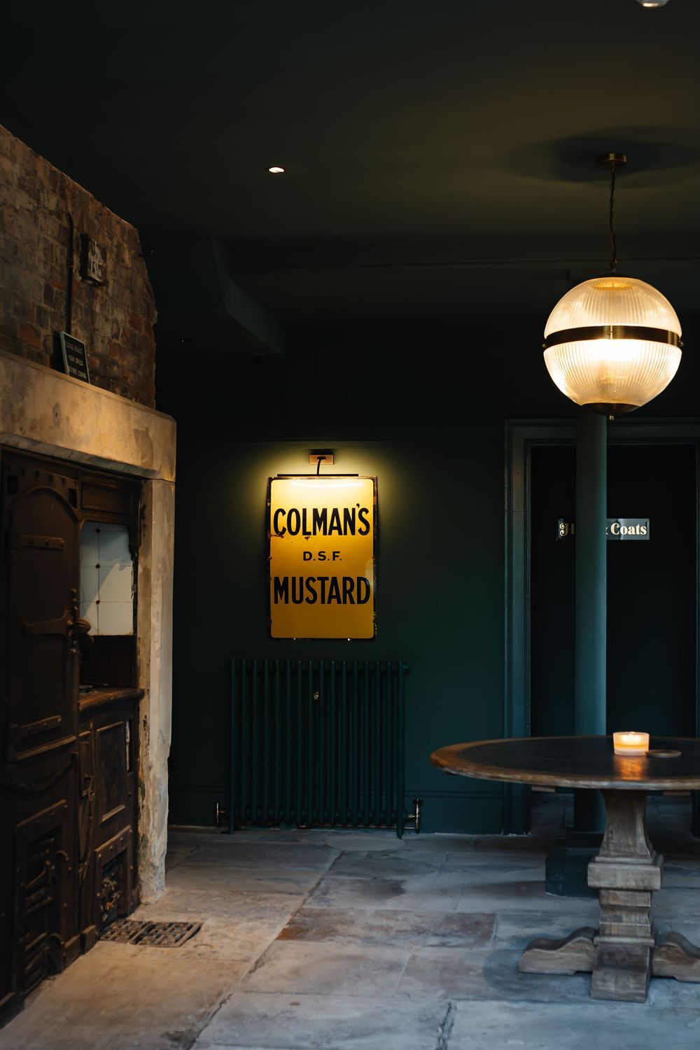 Vintage Colman's DSF Mustard illuminated sign on dark green wall above radiator in moody bar interior.