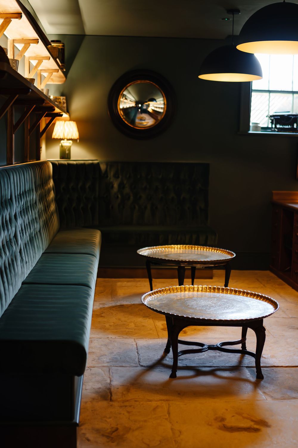 Dark moody lounge with tufted green velvet sofa, ornate brass coffee tables, round mirror, and pendant lamp.