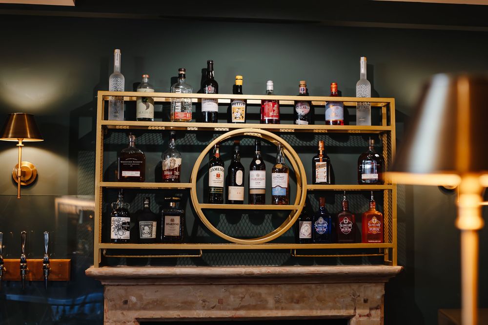 Gold bar shelf stocked with assorted liquor bottles against a dark green wall in a stylish bar.