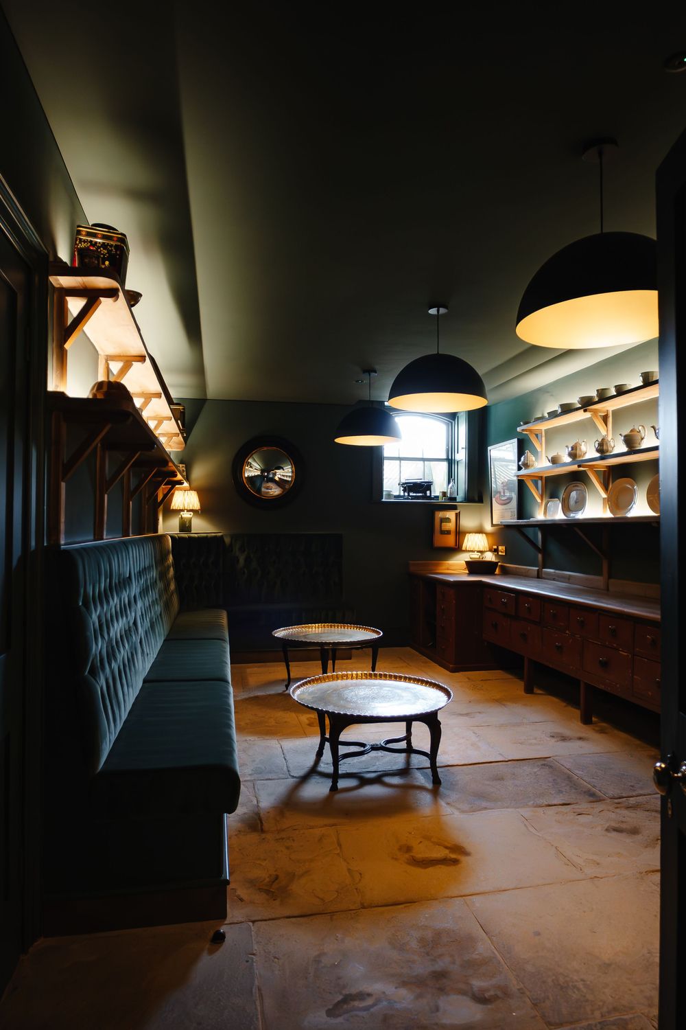 Dark moody lounge with tufted green sofa, round coffee tables, pendant lights, and rustic stone floor.